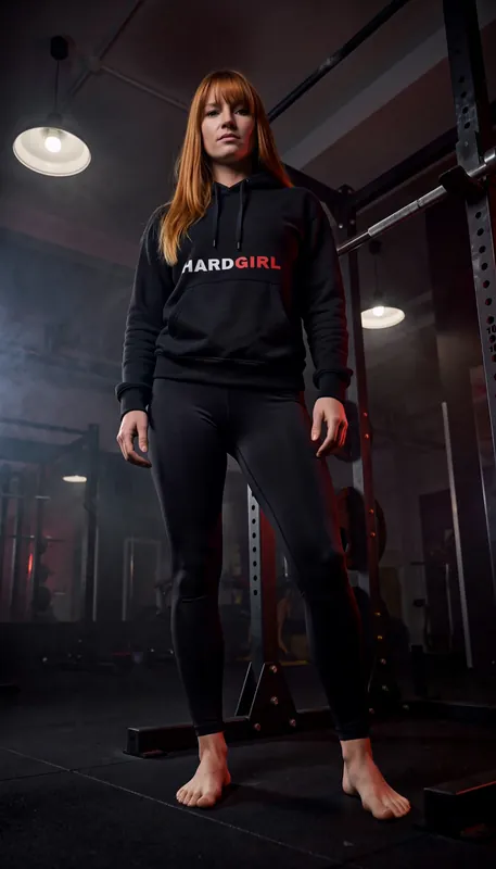 A red-haired woman in a black hoodie and leggings stands facing the camera in a dark gym.